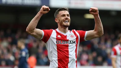A tight action shot of a muscular Oldham Athletic striker in a red and white striped jersey, mid-celebration after...