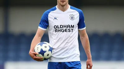 A photorealistic image of an Oldham Athletic player in a white shirt with blue sleeves and blue shorts, holding a...