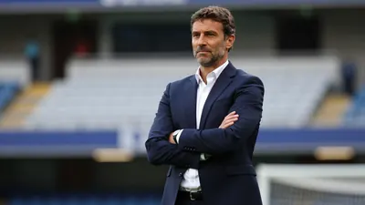 A 30-year-old football manager in a navy blue suit and white shirt, standing confidently with arms crossed in front of a...
