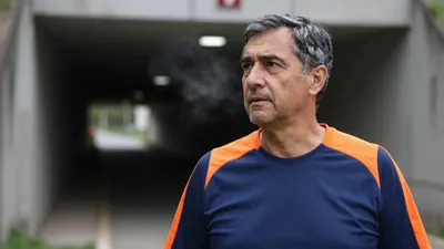 A 50-year-old male head coach in a plain blue and orange cotton training top, his face filled in a close medium shot as...