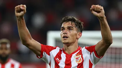 A young striker in a red and white striped kit, celebrating a goal with arms raised