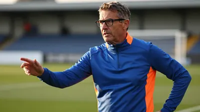 A close-up shot of a 45-year-old, middle-aged, bespectacled football manager in a plain blue and orange polyester...