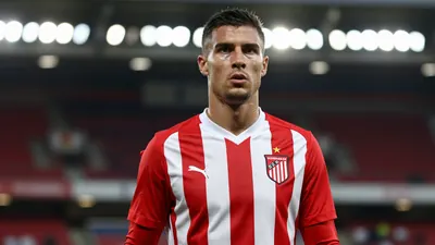 Photorealistic image of a 6'4" striker in a solid red and white striped kit, standing in a football stadium with a...