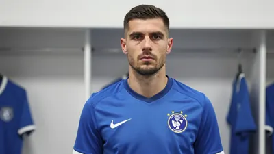 A 30-year-old defender in a solid blue football kit, standing in a dressing room with a focused expression, 85mm f/1.4...