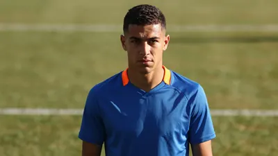 A determined 20-year-old midfielder with a lean build, wearing a plain blue and orange training top, intense eyes locked...