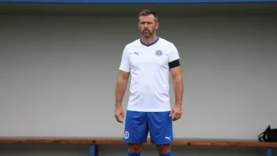A 35-year-old male coach with a strong build, wearing a white jersey with a blue collar and sleeves, a blue pair of...