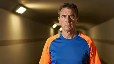 A 50-year-old male coach with a weathered complexion, standing in the dimly lit tunnel at Boundary Park, wearing a plain...