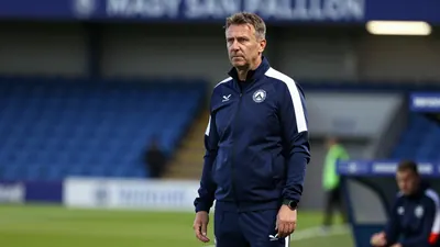 A 35-year-old Oldham Athletic coach, wearing a navy blue tracksuit with a white stripe, stands on the touchline at...