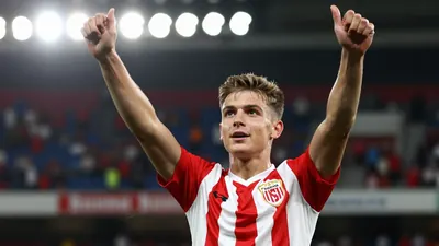 A 20-year-old forward in a red and white striped kit, celebrating a goal with arms raised, blurred stadium crowd in the...