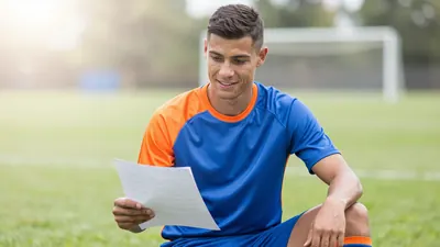Mid-20s male footballer, athletic build, waist-up, wearing a plain blue and orange training top (smooth polyester, solid...