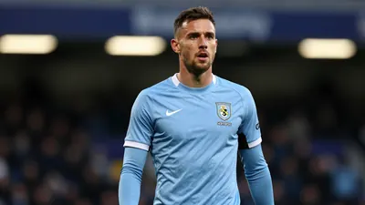 A mid-30s male Oldham Athletic player in a solid sky-blue kit, waist-up, focused expression, background blurred stadium...