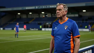 A 50-year-old Oldham coach, wearing a plain blue and orange cotton jersey, stands in the low-hued floodlight glow of...