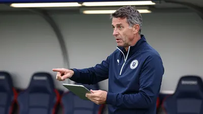 A mid-30s Oldham Athletic coach in a navy blue tracksuit with a white zip-up jacket, standing in a dugout, pointing at a...