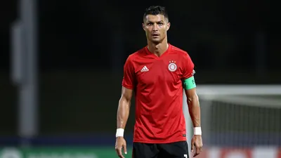 A 33-year-old midfielder with a determined expression, wearing a solid red shirt and black shorts, standing on a football...