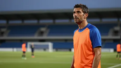 A 35-year-old, 5'9" male coach, dressed in a plain blue and orange polyester training top, stands in the deep bokeh of an...