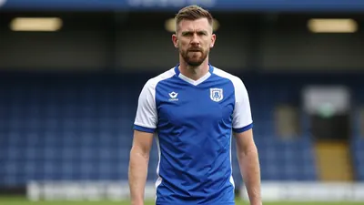 A photorealistic image of an anonymous Oldham Athletic AFC staff member, aged 30-40, standing in the Joe Royle Stand...