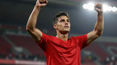 A young male striker in a red shirt, early 20s, muscular build, celebrating a goal with arms raised