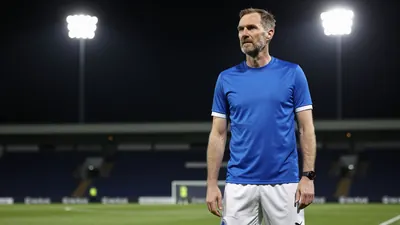 A photorealistic image of an anonymous Oldham Athletic coach, aged 30-40, standing on the 3G pitch, wearing a blue shirt...