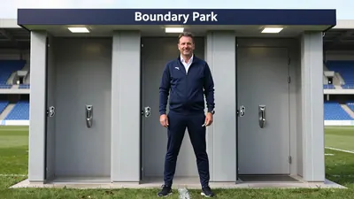 A 35-year-old stadium manager in a dark blue tracksuit, with a white shirt and a silver badge, stands in front of a newly...